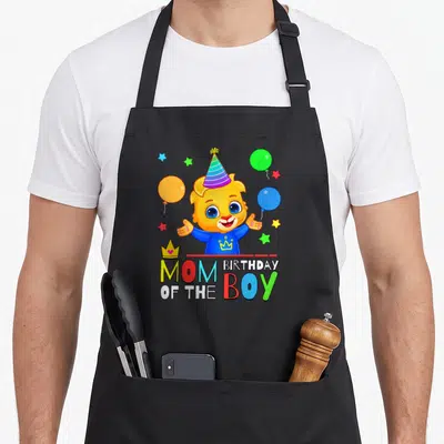 Lucas birthday apron - Cartoon lion cub in party hat with balloons, stars, crowns, and MOM OF THE BIRTHDAY BOY text — shown on a black cotton canvas apron against a white background