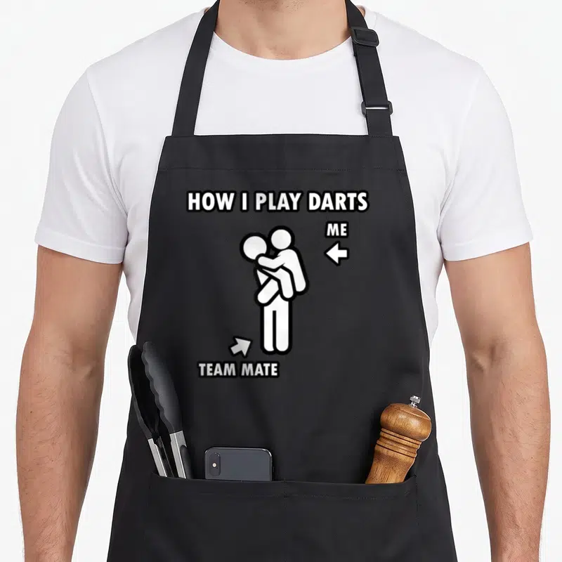 Dart friends apron - Text HOW I PLAY DARTS above two hugging stick figures labeled ME and TEAM MATE — shown on a black cotton canvas apron against a white background