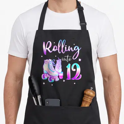 Rolling 12th birthday apron - Roller skates with Rolling into 12 text and stars in pink blue white — shown on a black cotton canvas apron against a white background