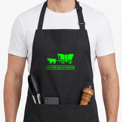 Died burpees apron - Green pixel art of a figure pulling a cart above a box reading YOU HAVE DIED OF BURPEES — shown on a black cotton canvas apron against a white background
