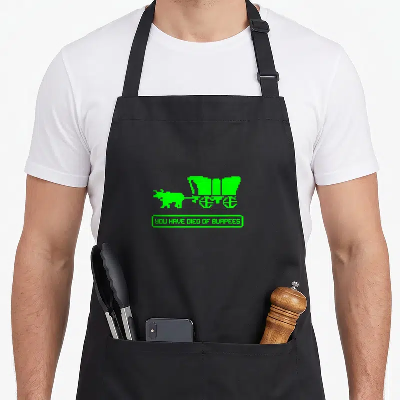 Died burpees apron - Green pixel art of a figure pulling a cart above a box reading YOU HAVE DIED OF BURPEES — shown on a black cotton canvas apron against a white background