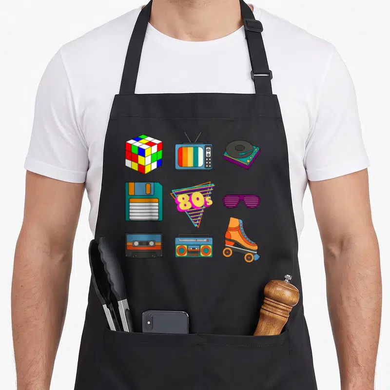 Throwback 80s apron - Cartoon grid of 80s items: Rubik's cube, TV, turntable, disk, glasses, boombox, skate, cassette — shown on a black cotton canvas apron against a white background