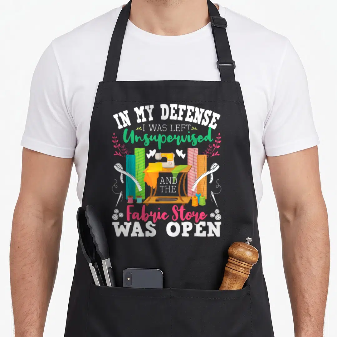 Unsupervised open apron - Cartoon sewing machine surrounded by colorful fabric rolls and measuring tapes with humorous — shown on a black cotton canvas apron against a white background