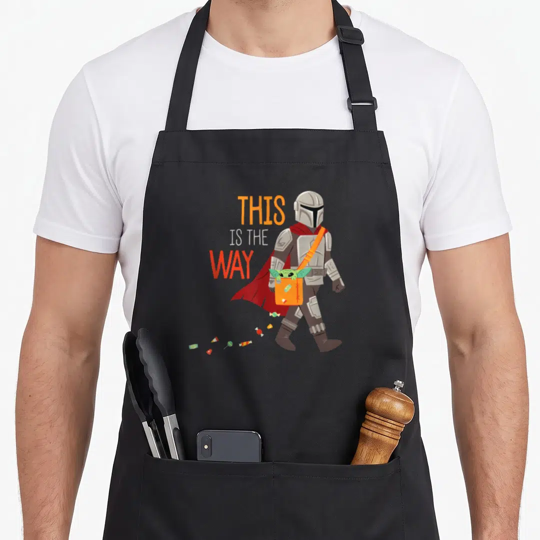 Way halloween apron - Mandalorian walking left with Grogu in bag and scattered candy. Stacked THIS IS THE WAY text — shown on a black cotton canvas apron against a white background