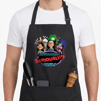 Jeff south boy apron - Neon Jeff Dunham text below a group of cartoon ventriloquist puppets and symbols including — shown on a black cotton canvas apron against a white background