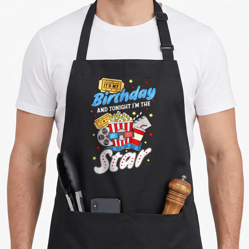Movie night family aprons - Birthday Star text with popcorn, tickets, 3D glasses, and film reel in blue red yellow — shown on a black cotton canvas apron against a white background