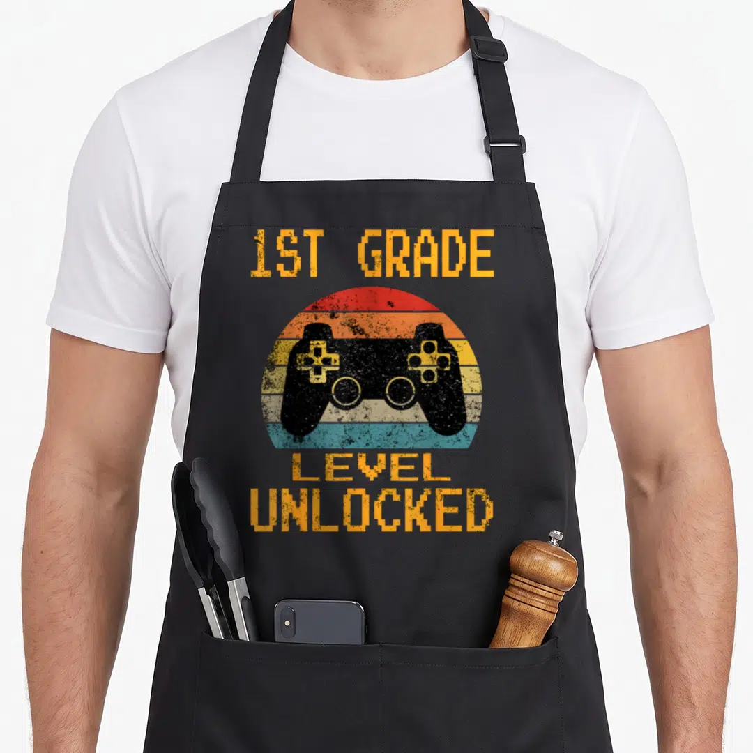 Grade gamer first apron - Retro game controller over orange and blue striped sunset with 1st grade level unlocked text — shown on a black cotton canvas apron against a white background
