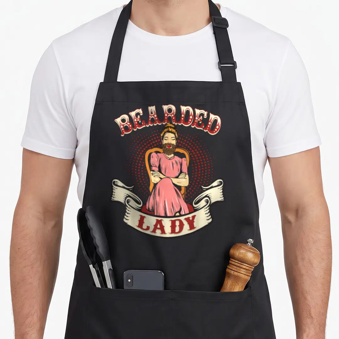 Circus bearded apron - Vintage woman with beard sitting in chair with BEARDED LADY text on red halftone background — shown on a black cotton canvas apron against a white background