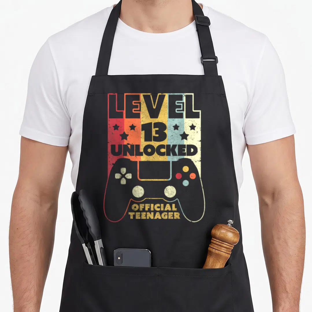 Level birthday apron - Retro game controller graphic with Level 13 Unlocked text and four stars on colored stripes — shown on a black cotton canvas apron against a white background