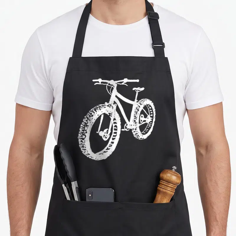 Tire fat man in apron - White line-art fat bike with wide knobby tires on black fabric — shown on a black cotton canvas apron against a white background