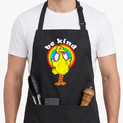 Kind bird apron - Yellow cartoon bird in front of rainbow circle under white be kind text — shown on a black cotton canvas apron against a white background