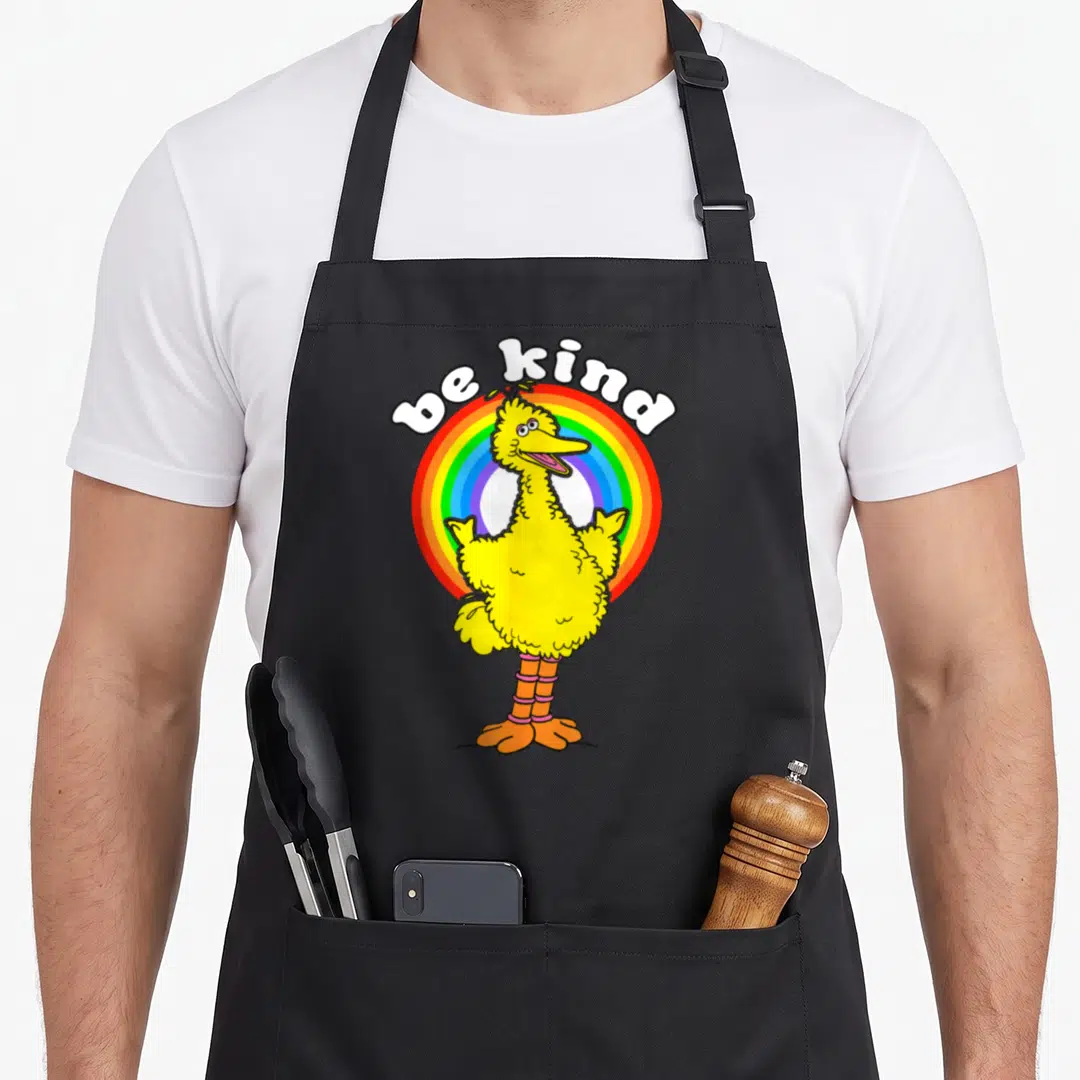 Kind bird apron - Yellow cartoon bird in front of rainbow circle under white be kind text — shown on a black cotton canvas apron against a white background