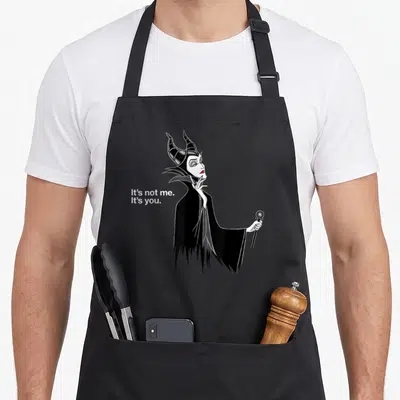 Villain novelty apron - Maleficent cartoon holding staff with text It's not me. It's you. in white — shown on a black cotton canvas apron against a white background