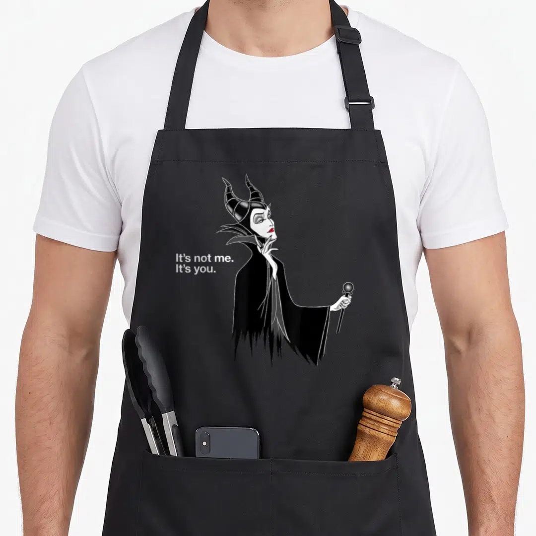 Villain novelty apron - Maleficent cartoon holding staff with text It's not me. It's you. in white — shown on a black cotton canvas apron against a white background