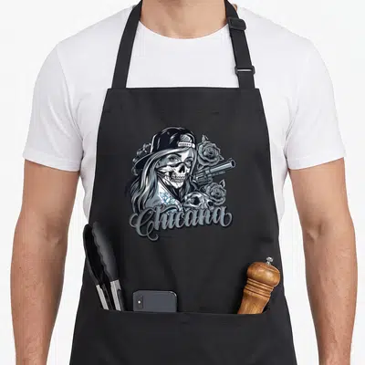 Chicana skull mexican apron - Woman in skull mask holding revolver with roses and Chicana script in black grey blue — shown on a black cotton canvas apron against a white background