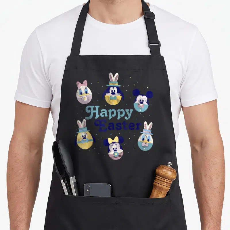 Funny friend easter egg apron - Five cartoon character eggs in blue yellow pink white surrounding centered Happy Easter — shown on a black cotton canvas apron against a white background