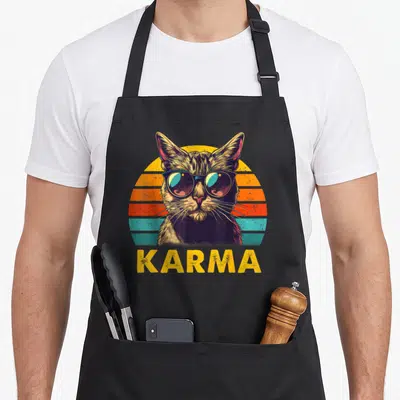 Music cat apron - Retro cat in sunglasses before striped sun with KARMA text in yellow orange red teal brown — shown on a black cotton canvas apron against a white background