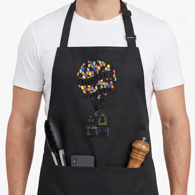 Adventure flying apron - House lifted by yellow blue pink orange balloons with two ribbons reading Adventure Is Out — shown on a black cotton canvas apron against a white background