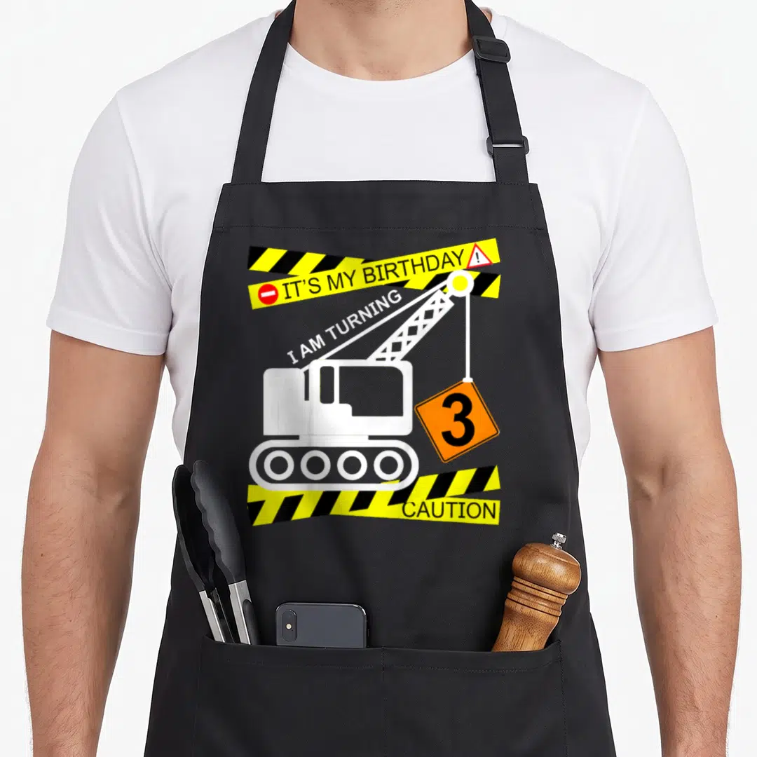Birthday construction truck apron - White excavator lifts orange sign with number 3 between yellow caution tape strips — shown on a black cotton canvas apron against a white background