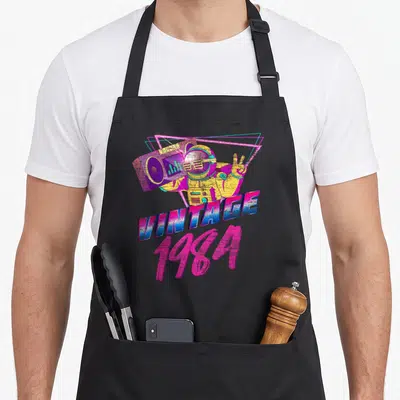 Sport 40th birthday apron - Retro astronaut holding purple boombox with neon triangles and VINTAGE 1984 text in pink — shown on a black cotton canvas apron against a white background