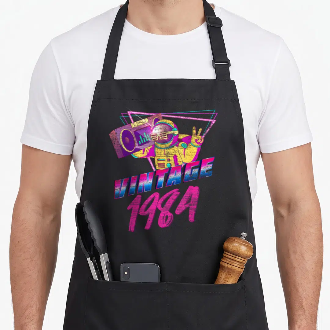 Sport 40th birthday apron - Retro astronaut holding purple boombox with neon triangles and VINTAGE 1984 text in pink — shown on a black cotton canvas apron against a white background