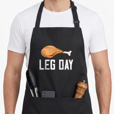 Leg costume thanksgiving day aprons - Orange cartoon chicken leg above white LEG DAY text — shown on a black cotton canvas apron against a white background
