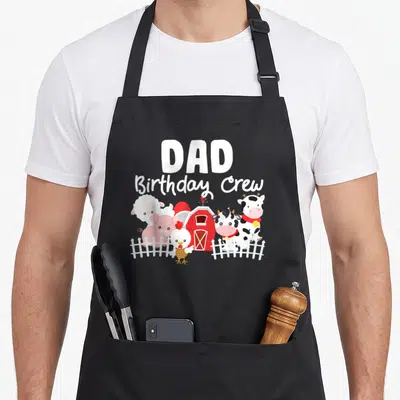 Birthday crew farm apron - DAD Birthday Crew text above cartoon cow pig sheep and chicken standing near red barn — shown on a black cotton canvas apron against a white background