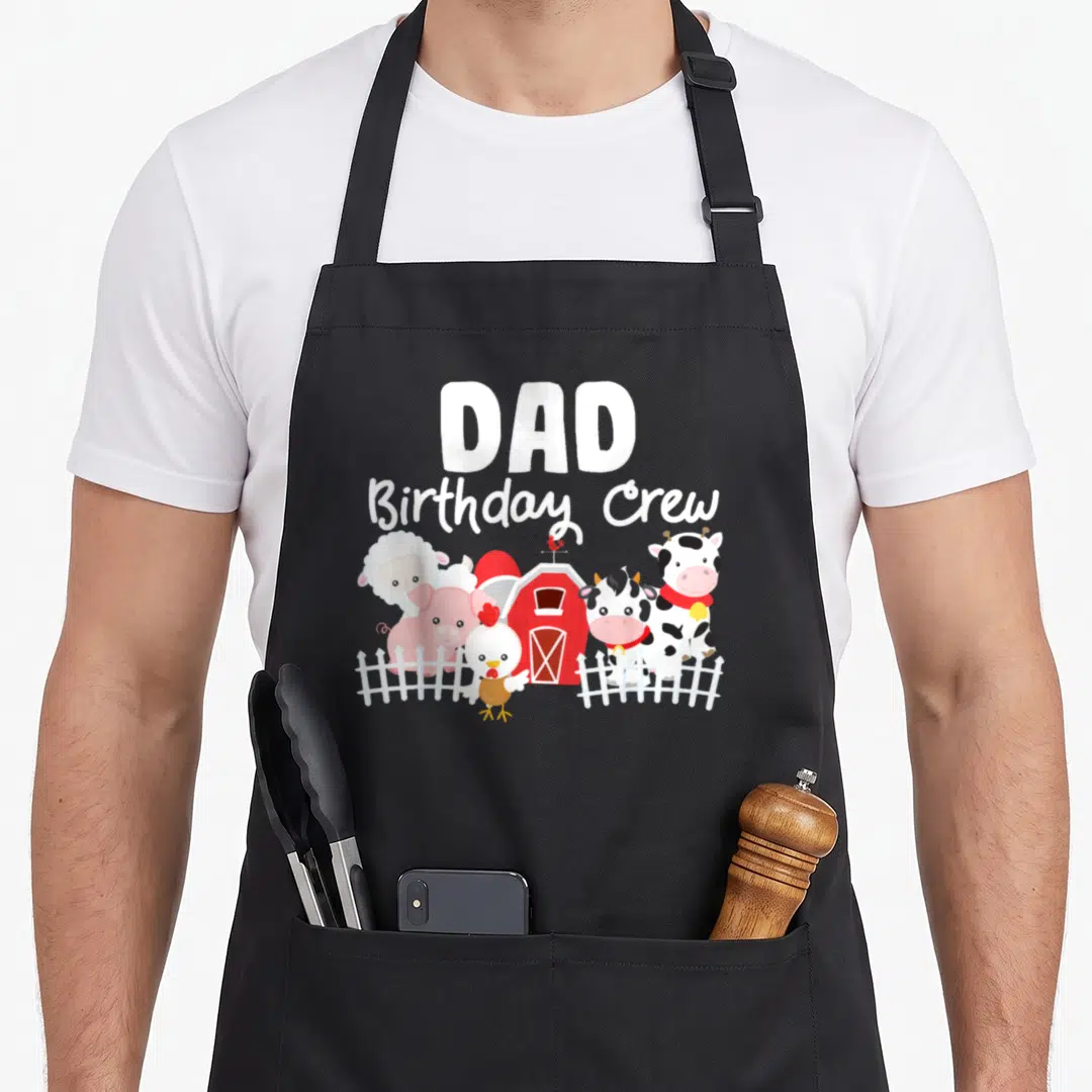 Birthday crew farm apron - DAD Birthday Crew text above cartoon cow pig sheep and chicken standing near red barn — shown on a black cotton canvas apron against a white background