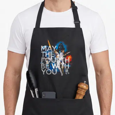Fourth may look apron - Stacked May The Fourth Be With You text over watercolor Star Wars character collage — shown on a black cotton canvas apron against a white background