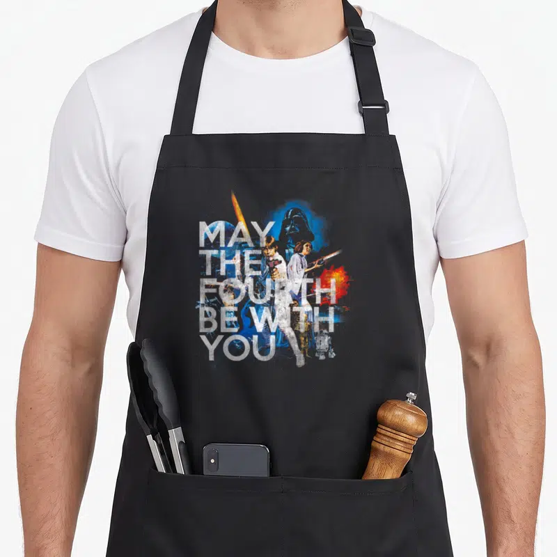 Fourth may look apron - Stacked May The Fourth Be With You text over watercolor Star Wars character collage — shown on a black cotton canvas apron against a white background
