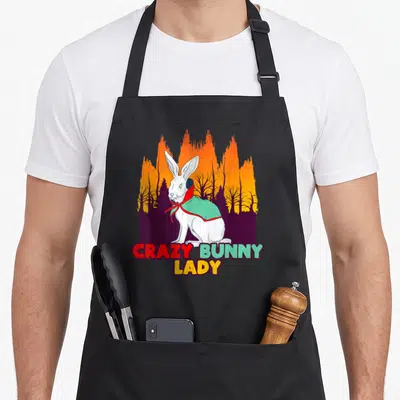 Rabbit halloween apron - White bunny in mint cape with CRAZY BUNNY LADY text over orange forest silhouette — shown on a black cotton canvas apron against a white background