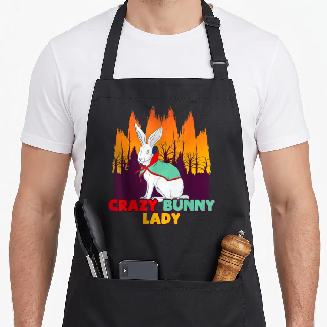 Rabbit halloween apron - White bunny in mint cape with CRAZY BUNNY LADY text over orange forest silhouette — shown on a black cotton canvas apron against a white background