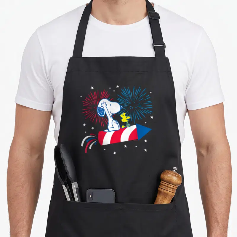 Woodstock 4th of july apron - Snoopy and yellow Woodstock sitting on a red and white striped rocket with blue nose — shown on a black cotton canvas apron against a white background