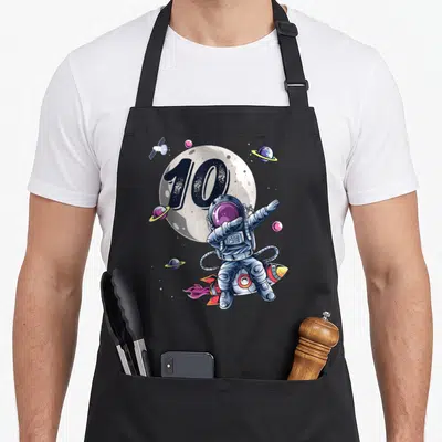 Space 10th birthday apron - Astronaut dabbing on rocket with 10 on moon and planets — shown on a black cotton canvas apron against a white background