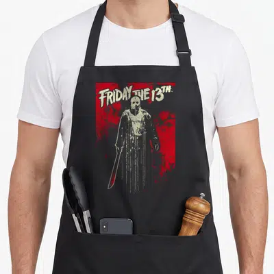 Jason drip apron - Arched FRIDAY THE 13TH text above figure holding machete on red splattered background — shown on a black cotton canvas apron against a white background