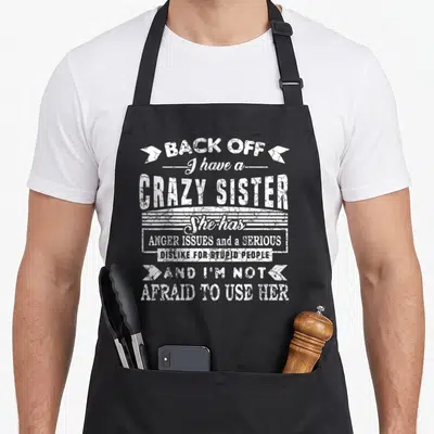 Sister use of apron - White distressed text reading Back Off I Have a Crazy Sister with arrows and horizontal lines — shown on a black cotton canvas apron against a white background
