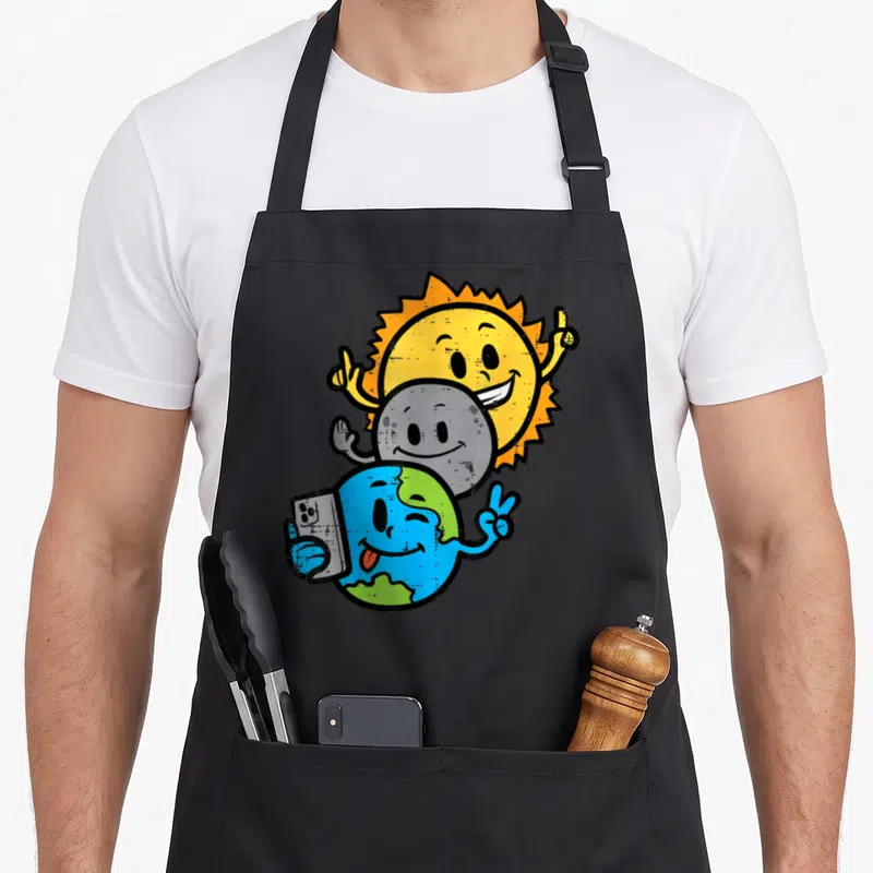 Selfie april apron - Cartoon Earth holding smartphone making peace sign with grey Moon and yellow Sun behind — shown on a black cotton canvas apron against a white background