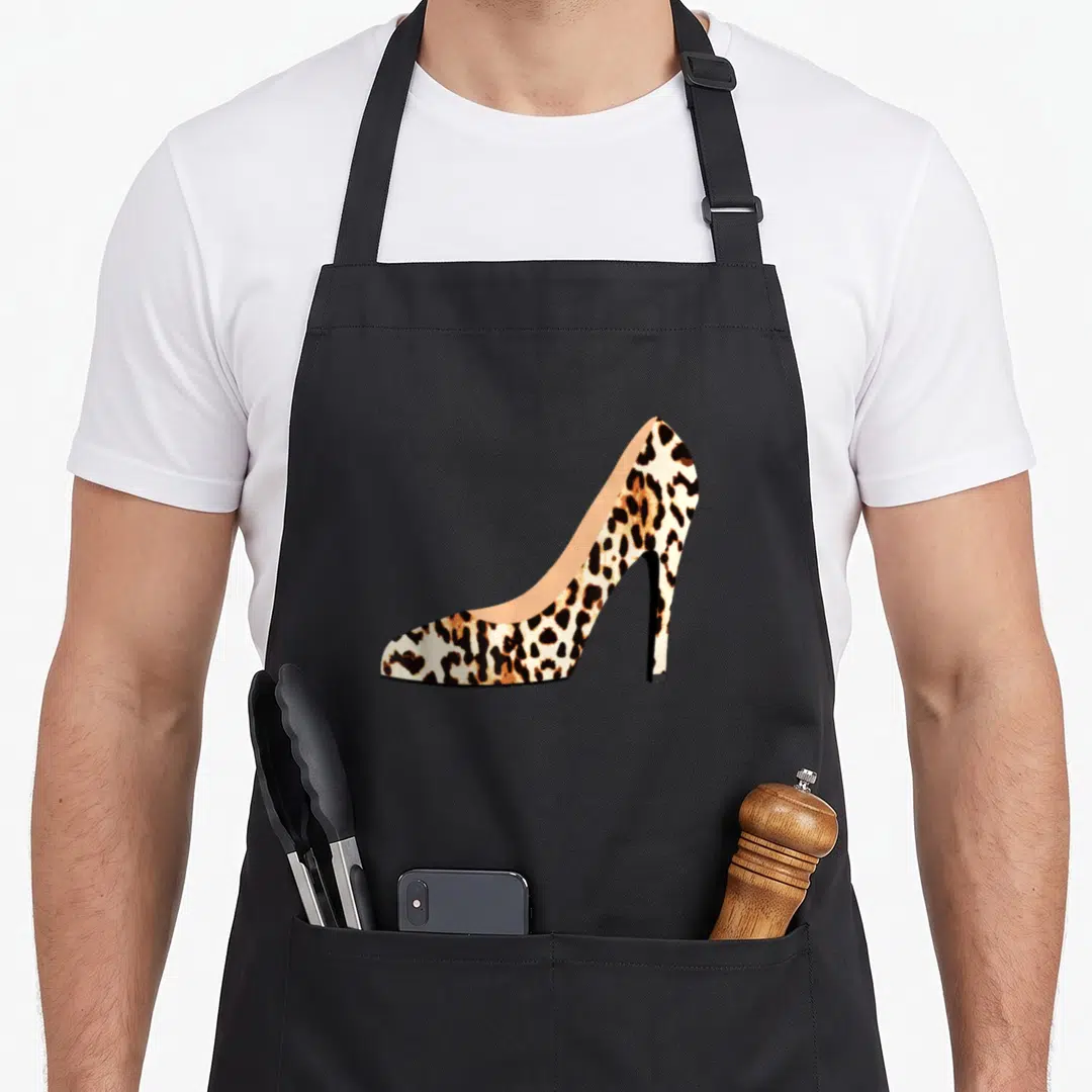 Heel leopard print apron - Realistic high heel shoe with tan brown and black leopard print pattern — shown on a black cotton canvas apron against a white background