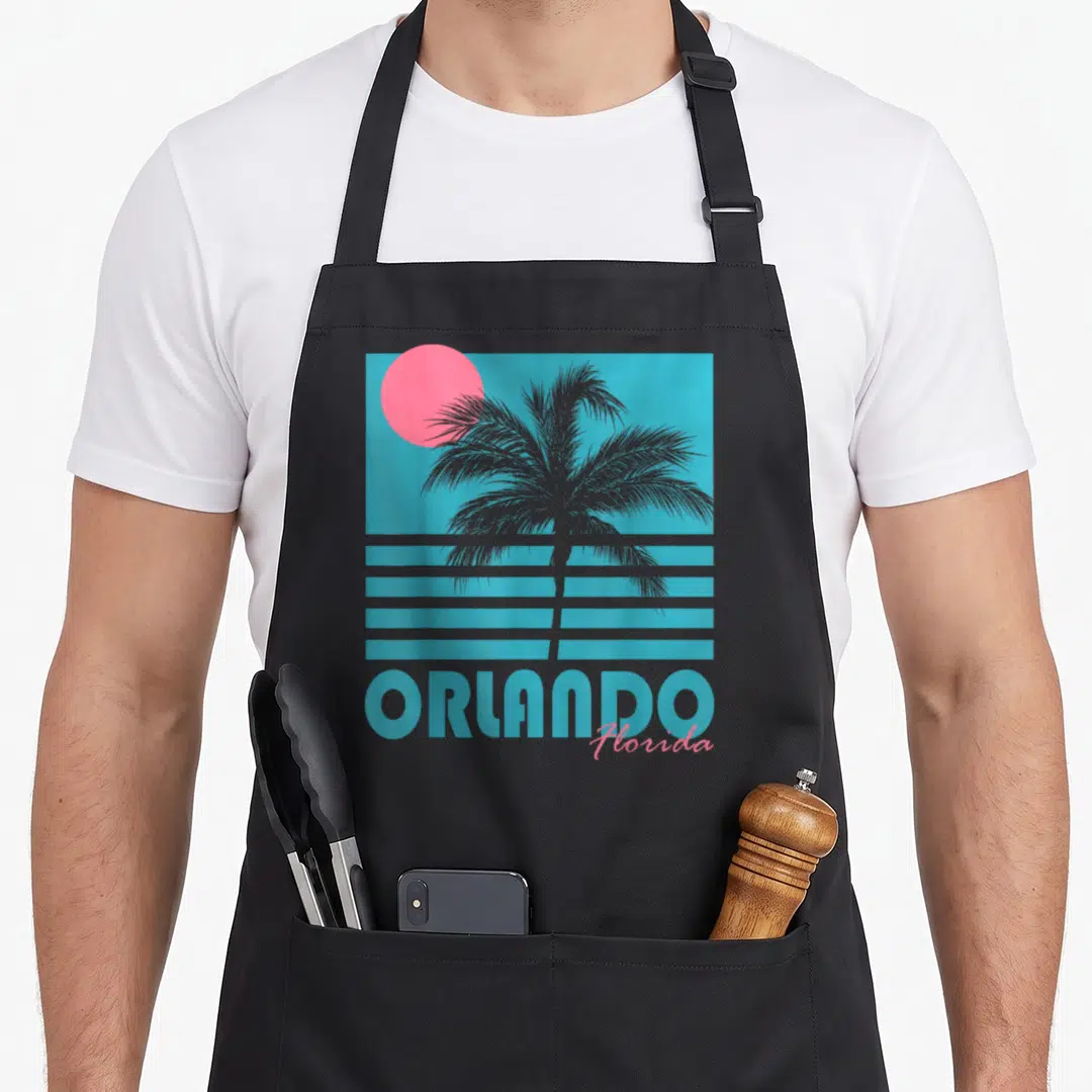 Orlando beach souvenir florida apron - Retro grey palm tree silhouette on blue background with pink sun and orange — shown on a black cotton canvas apron against a white background