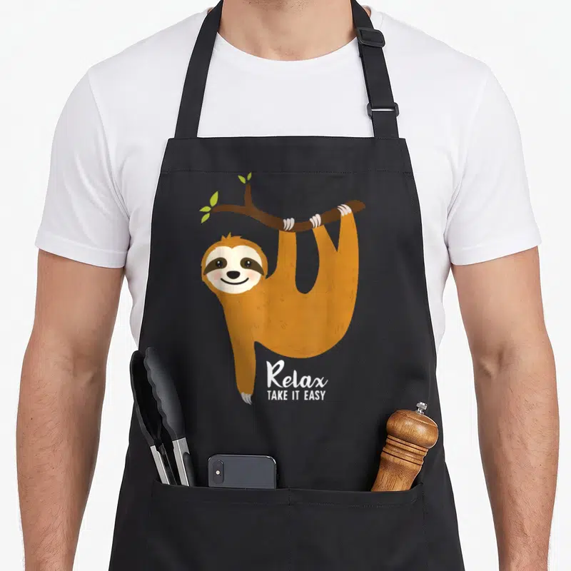 Hanging sloth apron - Brown cartoon sloth hanging from a branch with white Relax TAKE IT EASY text — shown on a black cotton canvas apron against a white background