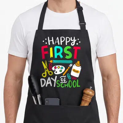 School art teacher apron - HAPPY FIRST DAY OF SCHOOL text with cartoon scissors, glue, pencil, and paint palette in red — shown on a black cotton canvas apron against a white background