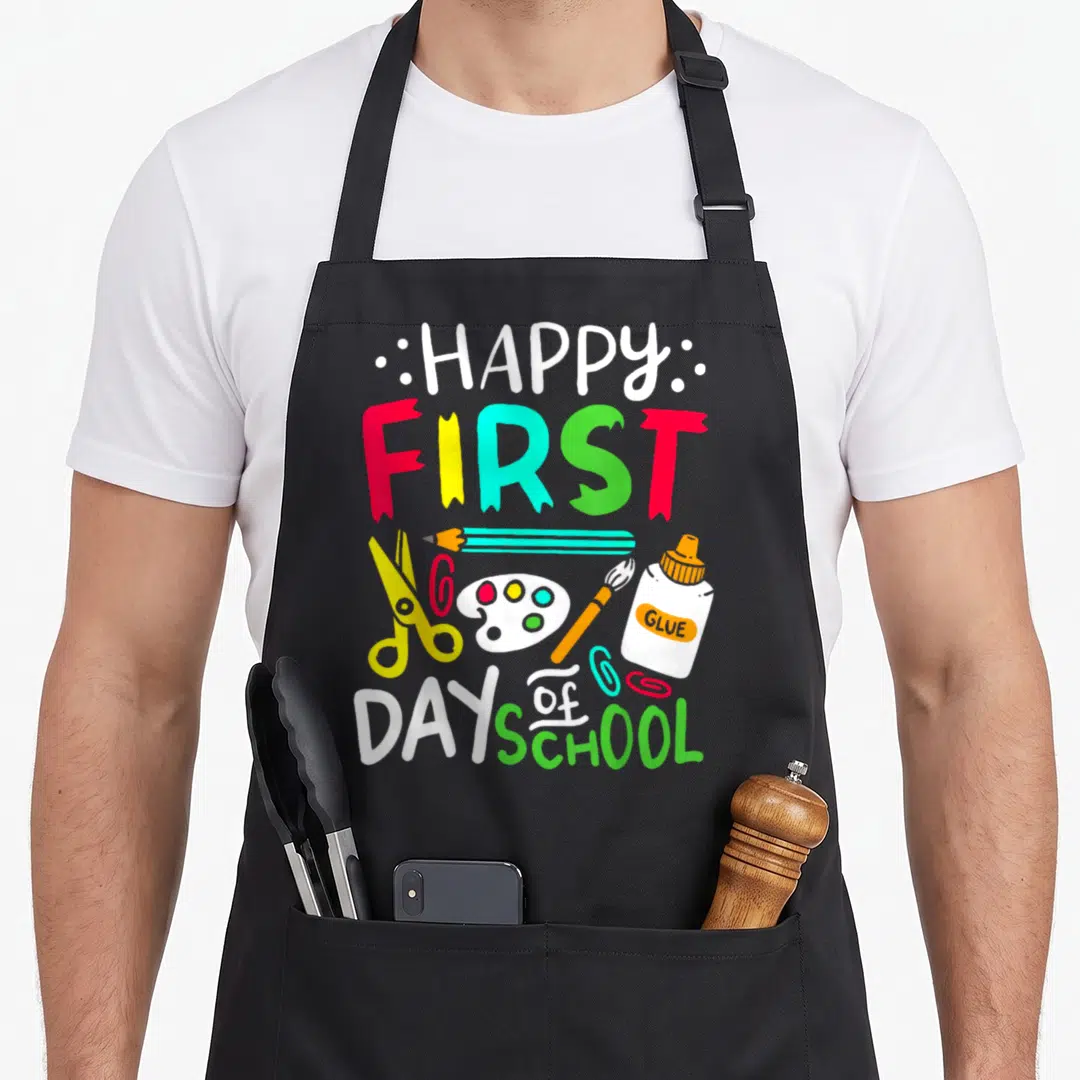 School art teacher apron - HAPPY FIRST DAY OF SCHOOL text with cartoon scissors, glue, pencil, and paint palette in red — shown on a black cotton canvas apron against a white background