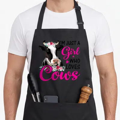 Cow farmer farm apron - Realistic cow head with pink flower crown and text I'M JUST A Girl WHO LOVES Cows — shown on a black cotton canvas apron against a white background