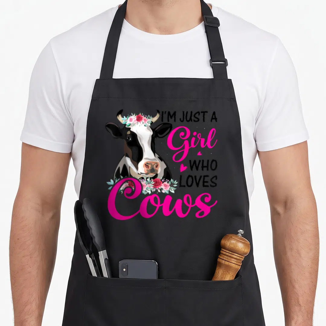 Cow farmer farm apron - Realistic cow head with pink flower crown and text I'M JUST A Girl WHO LOVES Cows — shown on a black cotton canvas apron against a white background