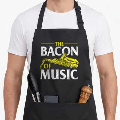 Saxophone bacon apron - Yellow distressed saxophone graphic between white stacked text reading The Bacon of Music — shown on a black cotton canvas apron against a white background