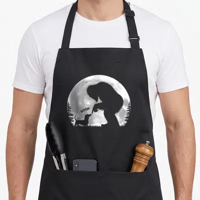 Hand dachshund apron - Bigfoot silhouette touching dachshund nose with BOOP text inside a white moon circle — shown on a black cotton canvas apron against a white background