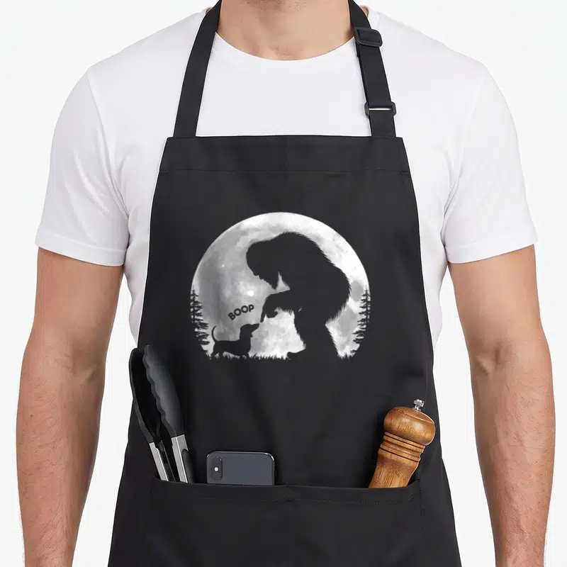 Hand dachshund apron - Bigfoot silhouette touching dachshund nose with BOOP text inside a white moon circle — shown on a black cotton canvas apron against a white background