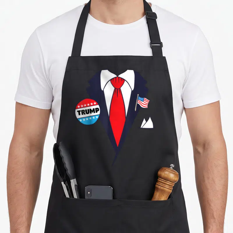 Donald cartoon apron - Navy suit red tie TRUMP button US flag white hair tuft cartoon print — shown on a black cotton canvas apron against a white background