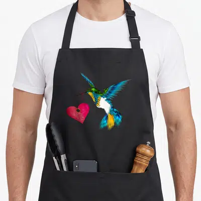 Cute hummingbird bird apron - Realistic hummingbird flying holding pink heart by red string in beak with colorful — shown on a black cotton canvas apron against a white background