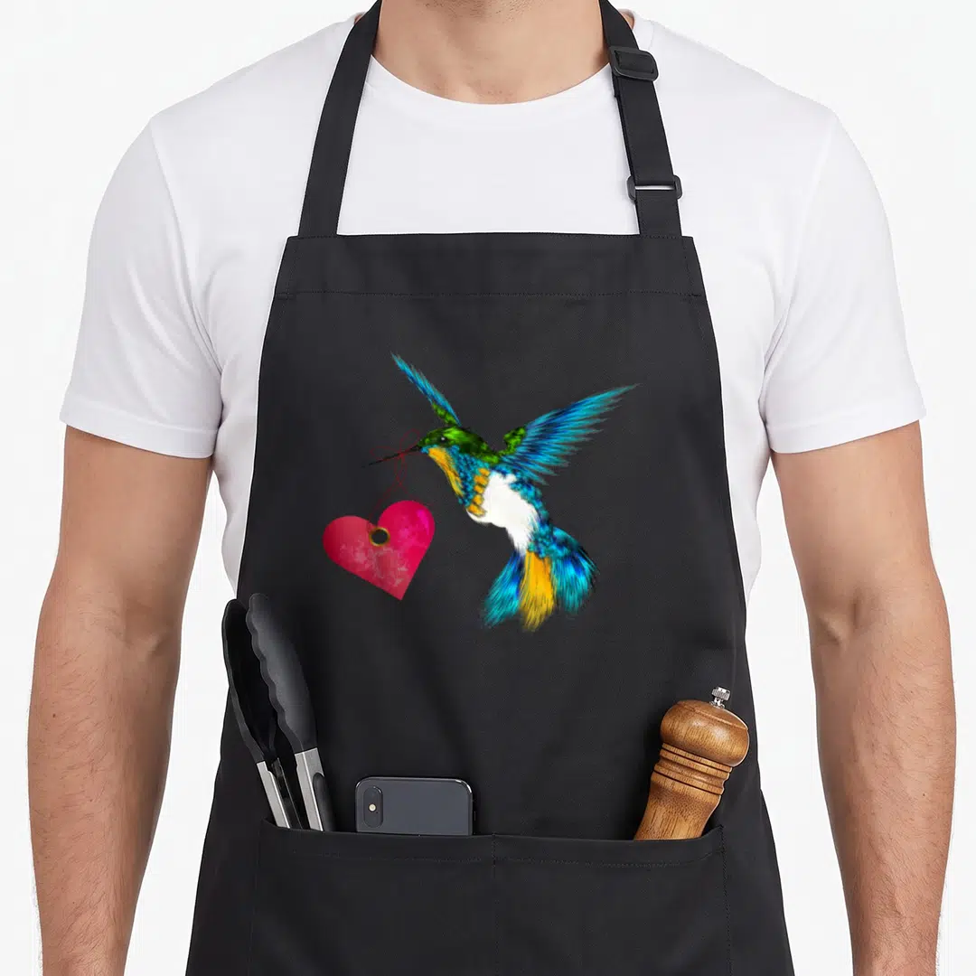 Cute hummingbird bird apron - Realistic hummingbird flying holding pink heart by red string in beak with colorful — shown on a black cotton canvas apron against a white background
